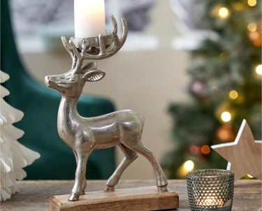 Silver reindeer candle holder on a wooden base with a lit pillar candle near a Christmas tree.